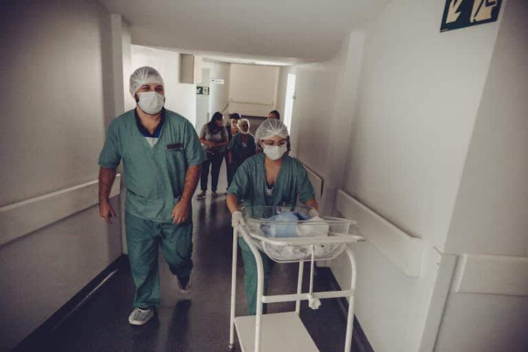 Healthcare workers in surgical attire transporting a newborn in a hospital corridor.